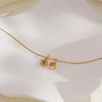El "Collar con inicial y foto", un delicado collar de oro con un disco redondo grabado con la letra K y un pequeño anillo de oro, capta maravillosamente el encanto de la joyería personalizada, expuesta con elegancia sobre una suave superficie blanca.