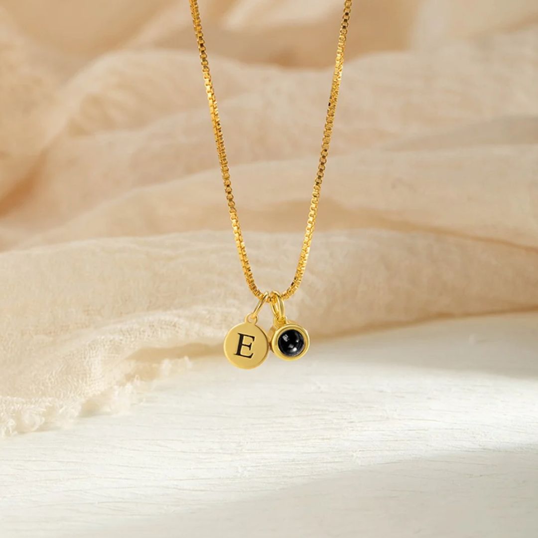 Un "Collar con inicial y foto" de oro con un colgante redondo con la letra E grabada y un pequeño colgante de piedra negra, sobre tela beige, ideal para quienes buscan collares personalizados con toques elegantes.