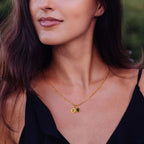Una mujer lleva un top negro sin mangas y el Collar con inicial y foto, un collar de oro con dos colgantes redondos grabados (E y M), sobre un fondo verde difuminado.