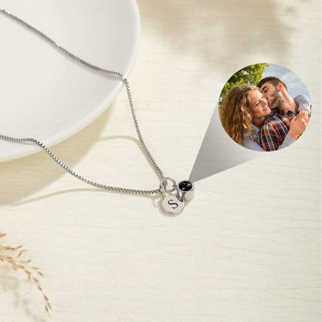 A "Collar con inicial y foto" de plata, con colgante en forma de letra S y un dije circular. Al hacer zoom, se revela una foto oculta en el interior. El collar está sobre una superficie clara.