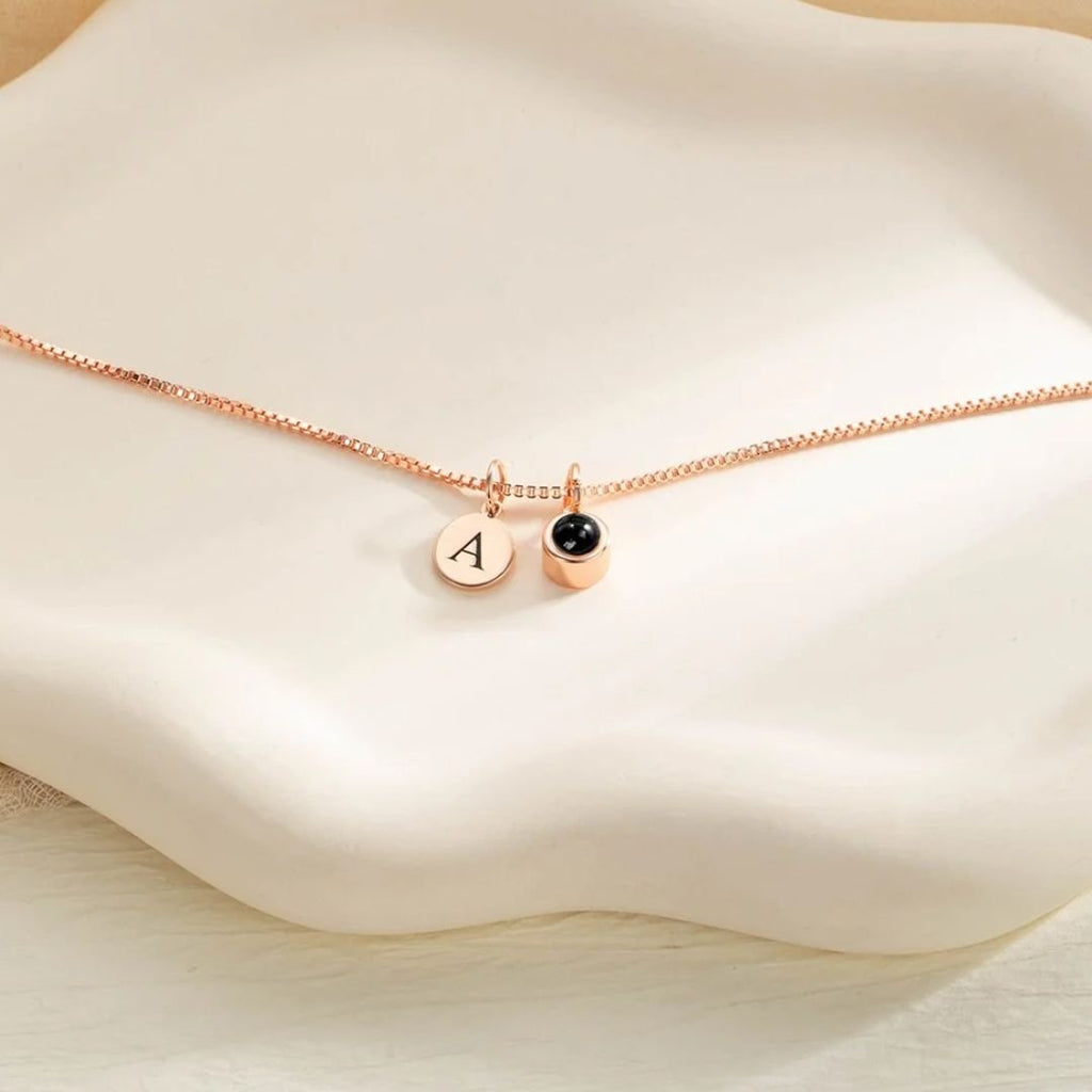 El Collar con inicial y foto es un collar en oro rosa con dos dijes: un disco con la letra A grabada y otro con piedra negra, expuesto sobre un plato cerámico blanco ondulado.