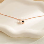 El Collar con inicial y foto es un collar en oro rosa con dos dijes: un disco con la letra A grabada y otro con piedra negra, expuesto sobre un plato cerámico blanco ondulado.