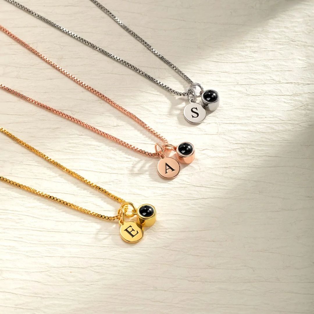 Tres collares con acabados en plata, oro rosa y oro, cada uno con un pequeño colgante redondo de cuenta negra y un colgante redondo grabado con una S, una A o una E, igual que el Collar con inicial y foto.