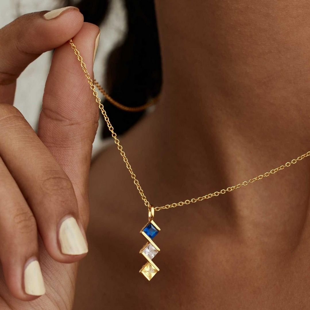 Una persona sostiene un Collar con múltiples piedras de nacimiento en oro personalizado, con tres gemas cuadradas-azul, transparente y amarilla-colgando en vertical. Se ven su piel y uñas claras. Regalo ideal para mamá.
