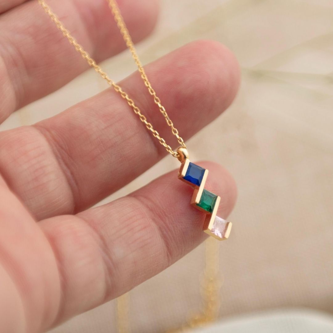Una mano sostiene el Collar con múltiples piedras de nacimiento, una cadena de oro con un colgante con tres piedras de nacimiento diagonales en azul, verde y rosa. Este collar personalizado es un regalo perfecto para mamá, con un fondo suavemente difuminado.