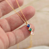Una mano sostiene el Collar con múltiples piedras de nacimiento, una cadena de oro con un colgante con tres piedras de nacimiento diagonales en azul, verde y rosa. Este collar personalizado es un regalo perfecto para mamá, con un fondo suavemente difuminado.