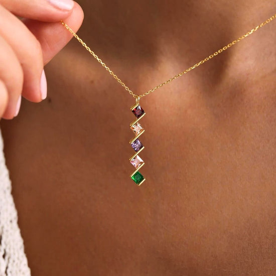 Primer plano de una persona que lleva el Collar con múltiples piedras de nacimiento en dorado, con un colgante vertical con cinco gemas cuadradas de colores. La mano sujeta la cadena cerca del cuello: un regalo ideal para mamá.