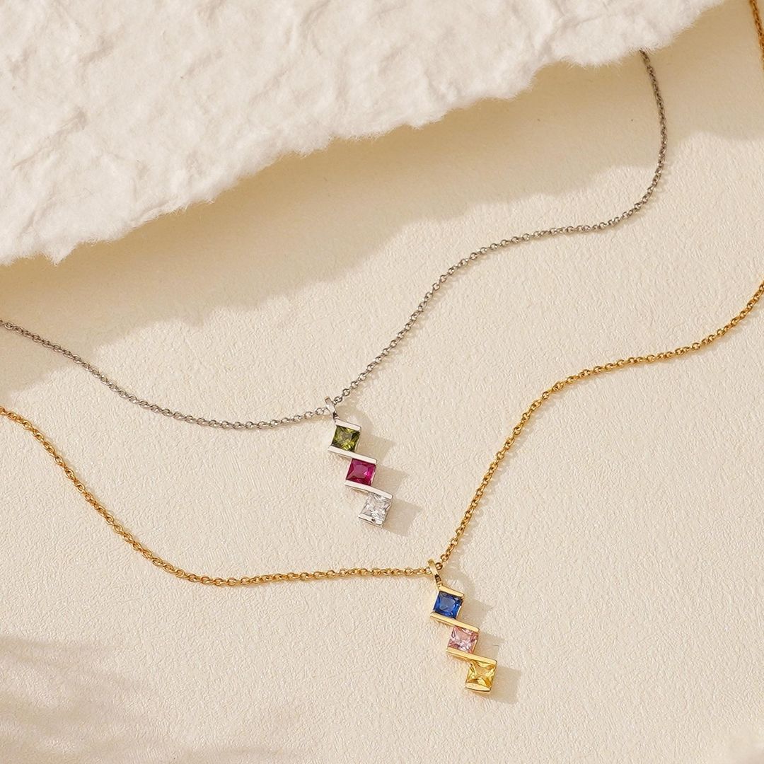 El Collar con múltiples piedras de nacimiento, disponible en oro y plata, muestra cuatro coloridas gemas de nacimiento; ideal como regalo para mamá y presentado sobre una superficie crema con tela suave en la esquina.