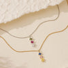 El Collar con múltiples piedras de nacimiento, disponible en oro y plata, muestra cuatro coloridas gemas de nacimiento; ideal como regalo para mamá y presentado sobre una superficie crema con tela suave en la esquina.
