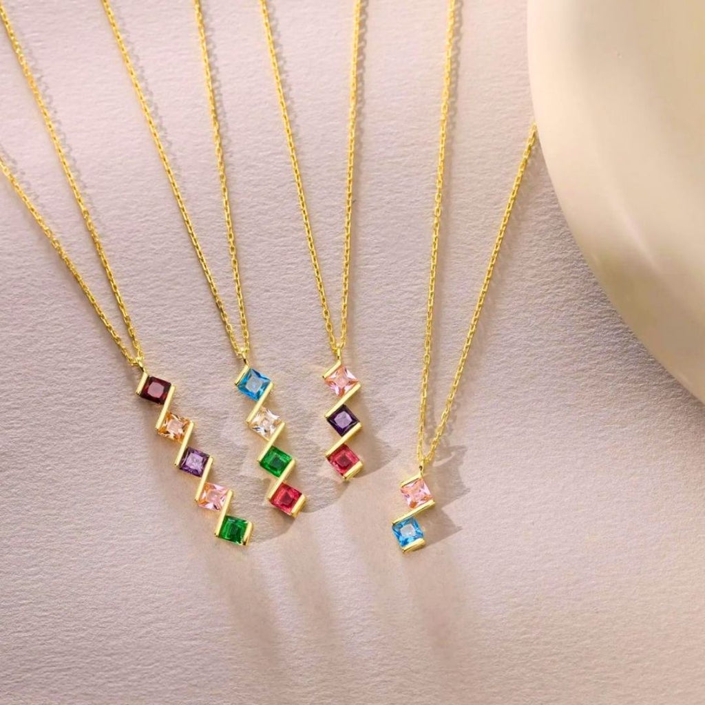 Cinco collares de oro, cada uno con un colgante rectangular de piedra de nacimiento en rojo, azul, verde, morado o rosa. El Collar con múltiples piedras de nacimiento es un regalo personalizado ideal para mamá.