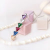 El Collar con piedras de nacimiento personalizadas presenta un dije vertical con seis gemas de colores en línea-amarillo, azul, rojo, verde, transparente y lila-ideal como regalo para mamá. Se muestra sobre fondo rosa suave y beige.