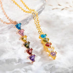 Dos collares "Collar con piedras de nacimiento personalizadas" de oro, con colgantes verticales que llevan seis piedras de diferentes formas y colores, ideales como regalo para mamá y exhibidos sobre mármol blanco.