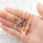 Una mano muestra cinco collares con piedras de nacimiento personalizadas, cada una de ellas con tres pequeñas gemas triangulares de colores engarzadas en oro o plata, dispuestas en fila sobre un fondo de textura clara.