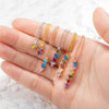 Una mano muestra cinco collares con piedras de nacimiento personalizadas, cada una de ellas con tres pequeñas gemas triangulares de colores engarzadas en oro o plata, dispuestas en fila sobre un fondo de textura clara.