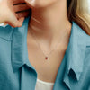Una mujer con camisa celeste lleva un Collar con piedras de nacimiento personalizadas con un pequeño dije rojo, apoyando la mano en su cara-el regalo perfecto para mamá.