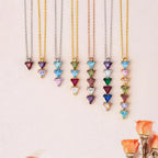 Nueve collares con piedras de nacimiento personalizadas se exhiben en fila sobre una superficie clara. Pequeñas rosas naranjas acentúan la parte inferior derecha, haciendo de estos collares un perfecto regalo para mamá o familiar de cuello.