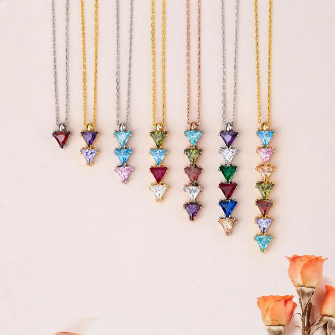 Nueve collares con piedras de nacimiento personalizadas se exhiben en fila sobre una superficie clara. Pequeñas rosas naranjas acentúan la parte inferior derecha, haciendo de estos collares un perfecto regalo para mamá o familiar de cuello.