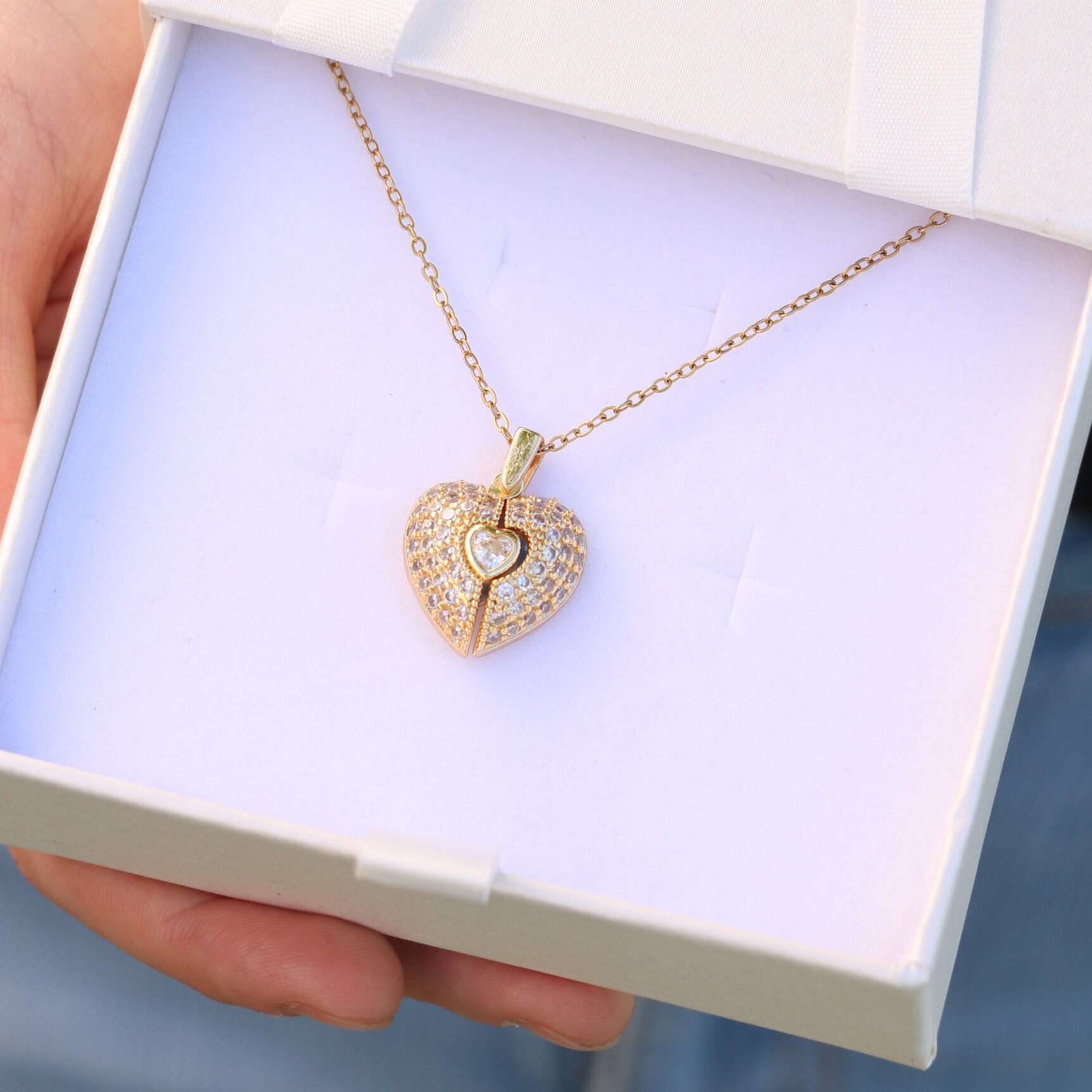 Una mano sostiene una caja blanca abierta que muestra el "Collar Corazón con alas de ángel y foto": un colgante dorado en forma de corazón con alas de ángel, un espacio para una foto y una delicada cadena.