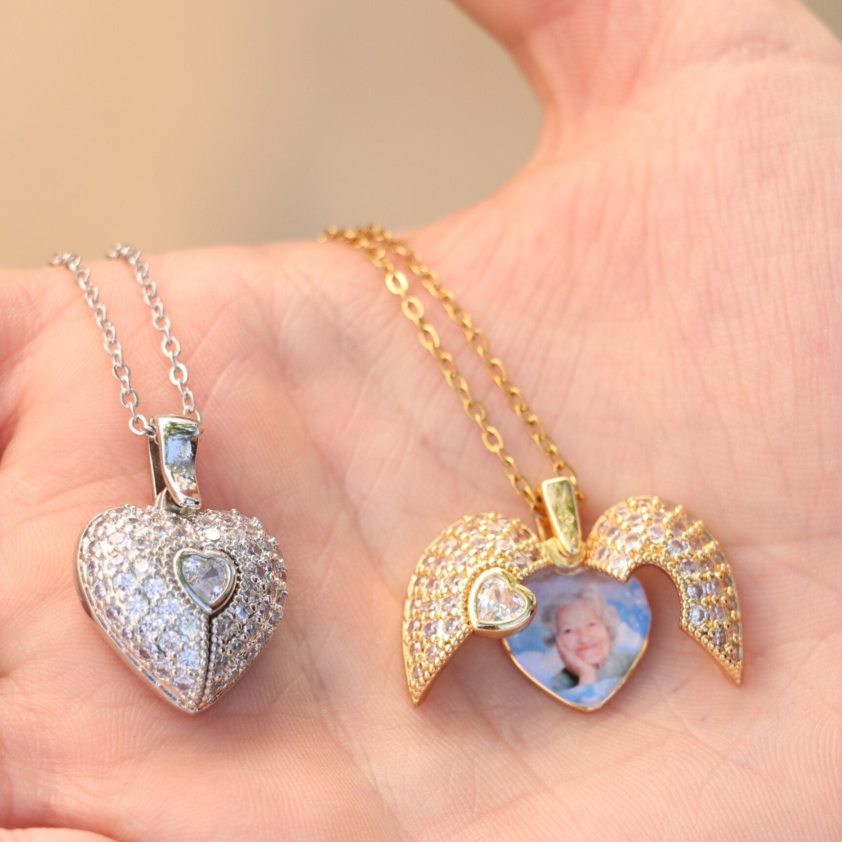Una mano sujeta dos colgantes en forma de corazón: un collar personalizado de plata y el collar de oro Corazón con alas de ángel y foto, con piedras brillantes, laterales en forma de alas y una foto en su interior.