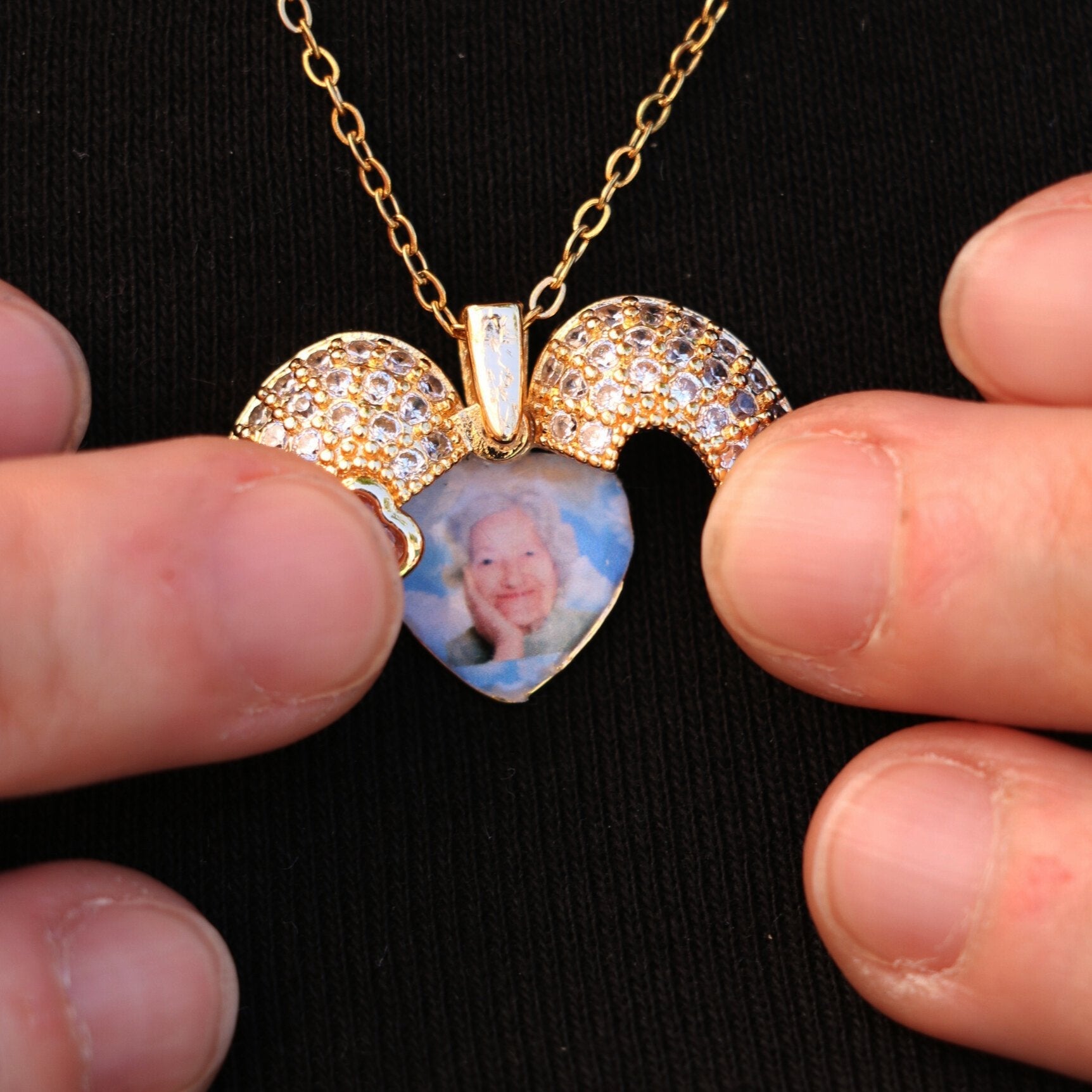 Primer plano de unas manos que sostienen un collar Corazón con alas de ángel y foto con un colgante en forma de corazón, detalles de pedrería y una foto de una anciana en su interior, sobre un fondo de tela negra.