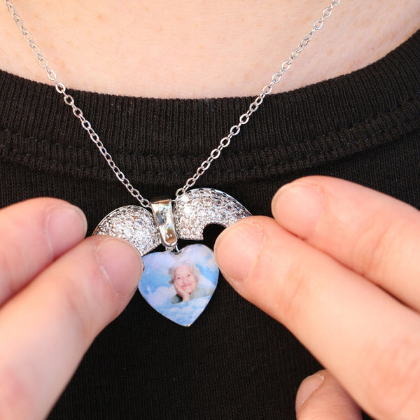 Una persona sostiene el Collar Corazón con alas de ángel y foto, que contiene la imagen de una mujer mayor en su interior, sobre una camisa negra.
