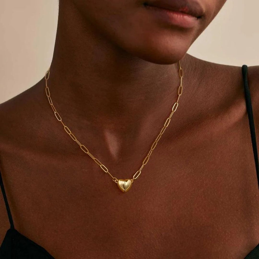 Una persona lleva el Collar Corazón magnético, un collar de cadena dorada con un colgante en forma de corazón, combinado con un top negro de tirantes. Se muestra desde los hombros hasta la barbilla. Ideal como regalo de San Valentín o collar de pareja.