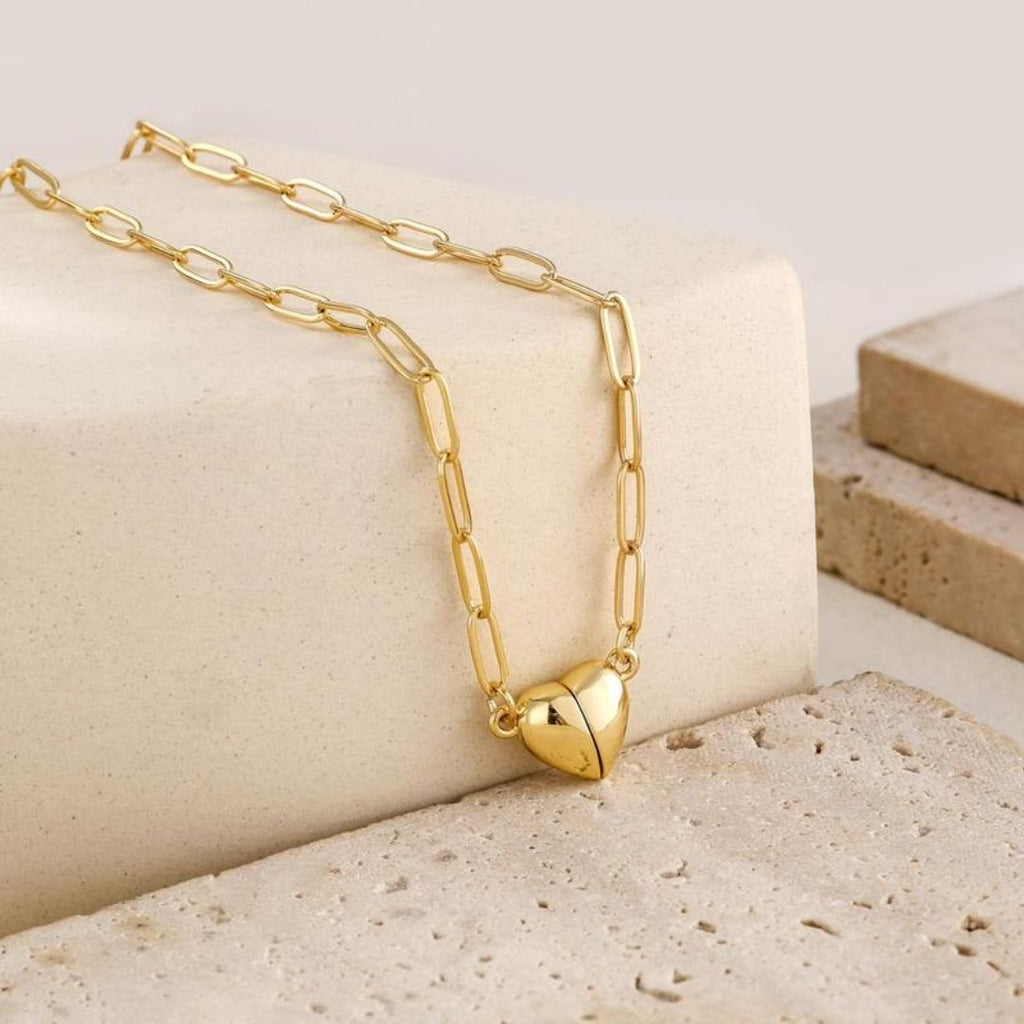 El collar Corazón magnético, un collar de cadena de oro con eslabones alargados y un colgante de corazón abullonado, se muestra sobre piedra beige, perfecto como collar de pareja o regalo de San Valentín para su amada.