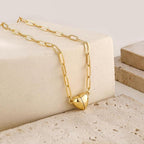 El collar Corazón magnético, un collar de cadena de oro con eslabones alargados y un colgante de corazón abullonado, se muestra sobre piedra beige, perfecto como collar de pareja o regalo de San Valentín para su amada.