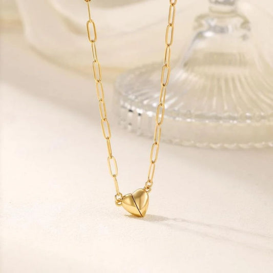 El collar Corazón magnético presenta eslabones alargados de oro y un colgante de corazón facetado, ideal como collar de pareja o regalo de San Valentín. Se muestra sobre un fondo neutro con un adorno de cristal en la distancia.