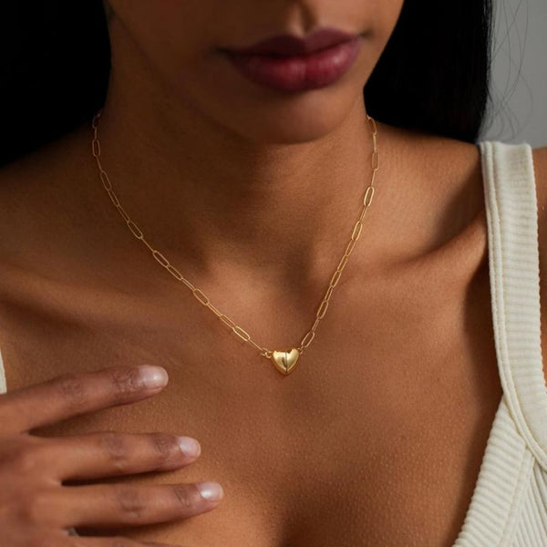 Una mujer luce el Collar Corazón magnético, un collar dorado con dije de corazón, combinando con una blusa blanca. Su mano cerca del cuello resalta el collar, perfecto como regalo de San Valentín o collar de pareja.