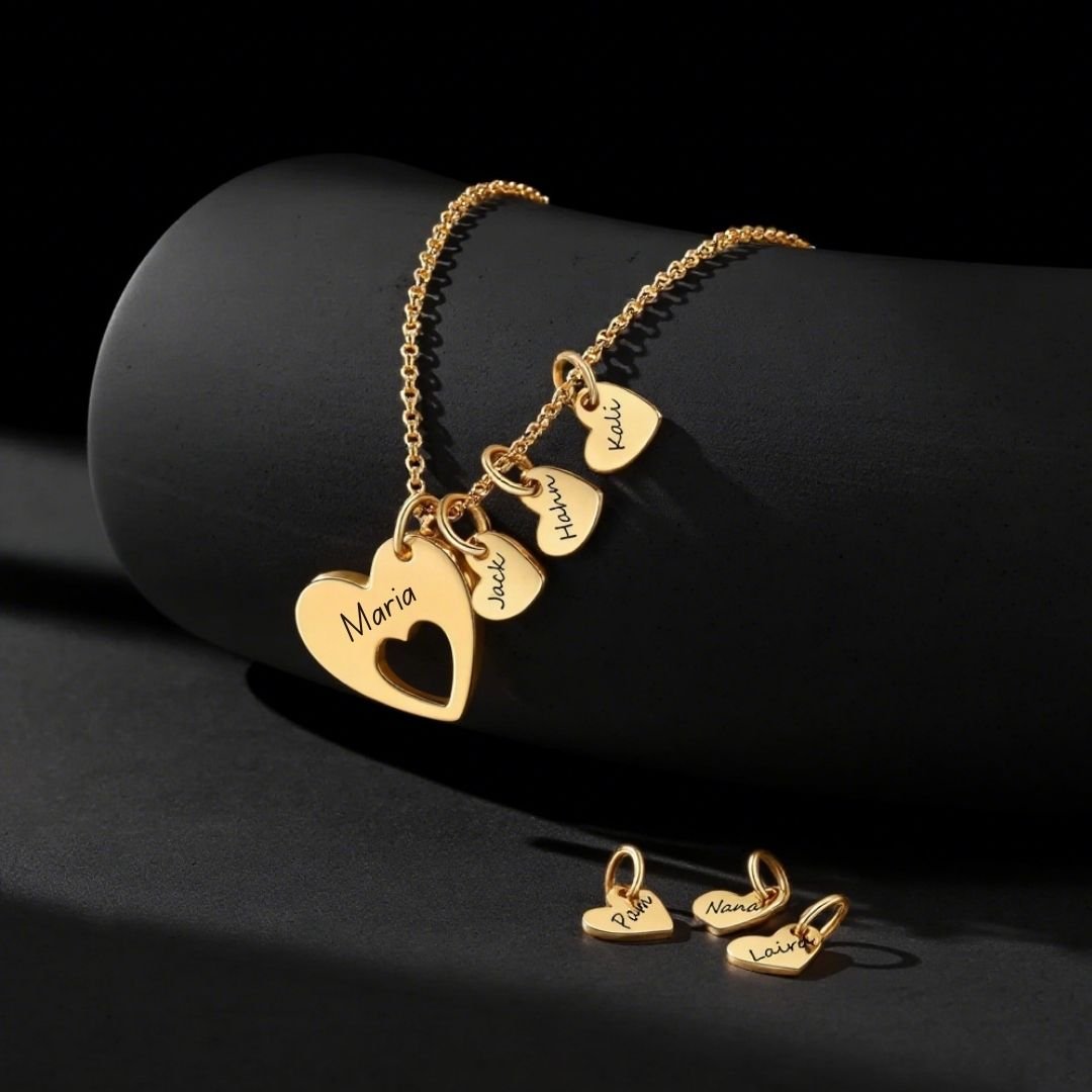 El collar Corazones de familia presenta colgantes de oro en forma de corazón grabados con los nombres de María, Julia, Hubert y Lukas, además de pequeños colgantes de oro en forma de corazón sobre una superficie negra.