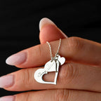 Una mano con las uñas cuidadas sostiene el collar Corazones de familia, un collar de plata con dos colgantes en forma de corazón -uno abierto y otro con la inscripción "Say Alice"- sobre un fondo negro.