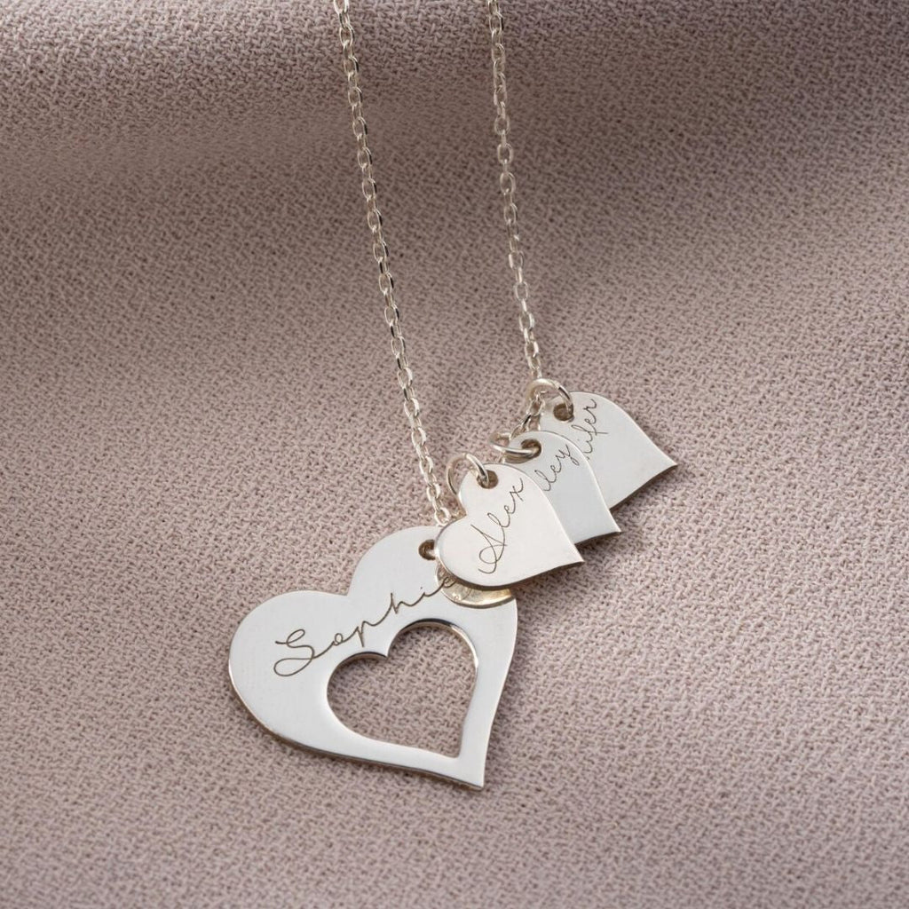 El collar Corazones de familia, un collar de plata con tres colgantes en forma de corazón grabados con Sophia, Olivia y Ava, se exhibe sobre una superficie beige texturizada: un elegante regalo personalizado para los seres queridos.
