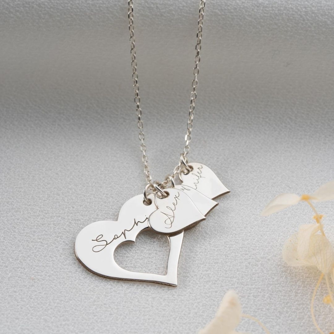 El collar Corazones de familia es un collar de plata con tres colgantes en forma de corazón de distintos tamaños, cada uno de ellos grabado con los nombres Sophia, Avery y Olivia en cursiva, sobre un fondo de tela gris claro.