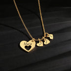 El collar Corazones de familia presenta una cadena de oro con un gran colgante de corazón grabado con "María" y tres corazones más pequeños etiquetados con "Jack", "Hana" y "Tú", expuestos sobre una superficie oscura y texturizada. Ideal como regalo personalizado.