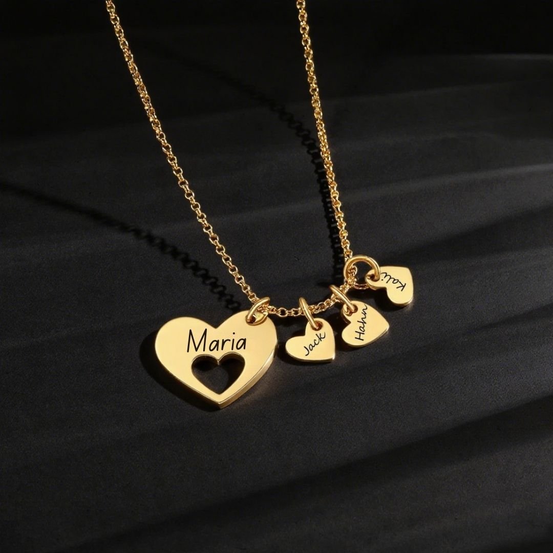 El collar Corazones de familia presenta una cadena de oro con un gran colgante de corazón grabado con "María" y tres corazones más pequeños etiquetados con "Jack", "Hana" y "Tú", expuestos sobre una superficie oscura y texturizada. Ideal como regalo personalizado.