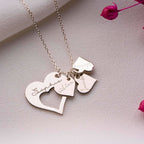 El collar Corazones de familia es un collar de plata con colgantes en forma de corazón grabados con los nombres Sophie, Aiden y Mummy, que se muestran sobre una superficie texturizada con flores secas de color rosa.