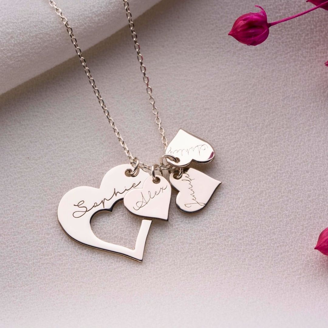 El collar Corazones de familia es un collar de plata con colgantes en forma de corazón grabados con los nombres Sophie, Aiden y Mummy, que se muestran sobre una superficie texturizada con flores secas de color rosa.