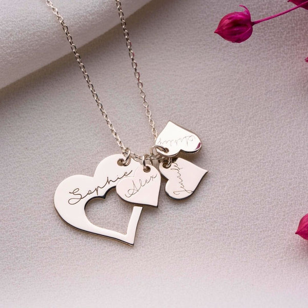 El collar Corazones de familia es un collar de plata con colgantes en forma de corazón grabados con los nombres Sophie, Aiden y Mummy, que se muestran sobre una superficie texturizada con flores secas de color rosa.