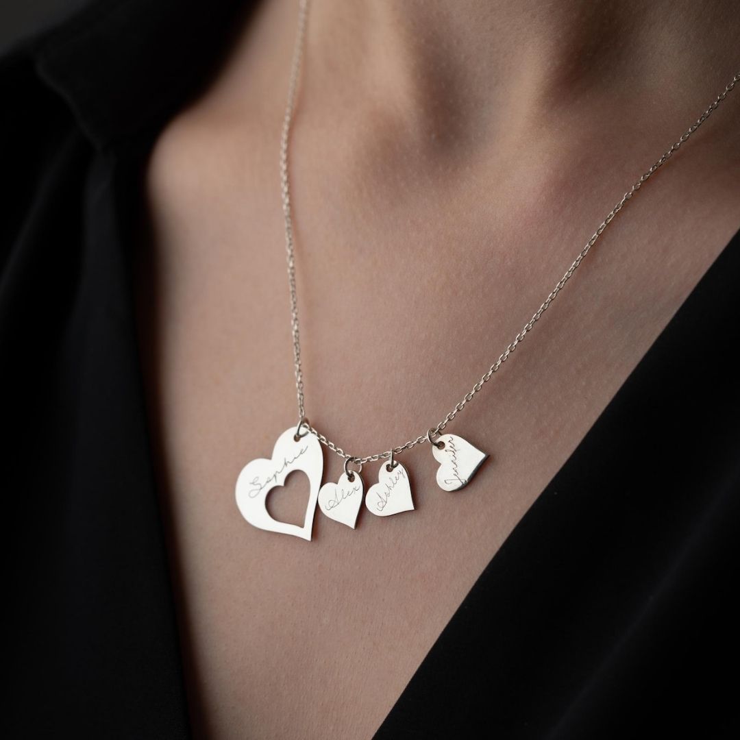 Primer plano de una persona que lleva el collar Corazones de familia, un collar de plata con cuatro colgantes en forma de corazón grabados con nombres, sobre un top negro de cuello de pico.