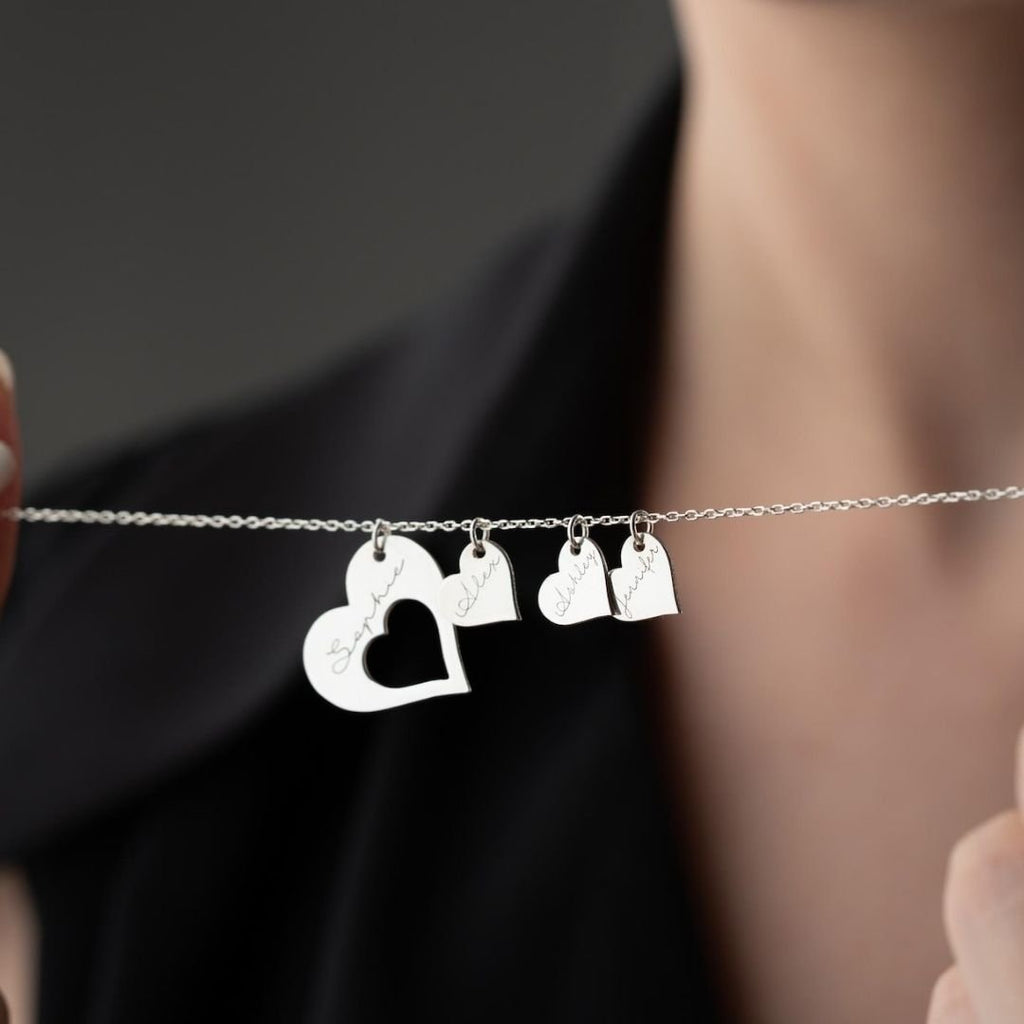 Una persona sostiene un collar Corazones de familia con cuatro dijes grabados: un corazón grande y tres formas más pequeñas con inscripciones en cursiva. El fondo está difuminado y la persona viste de negro.