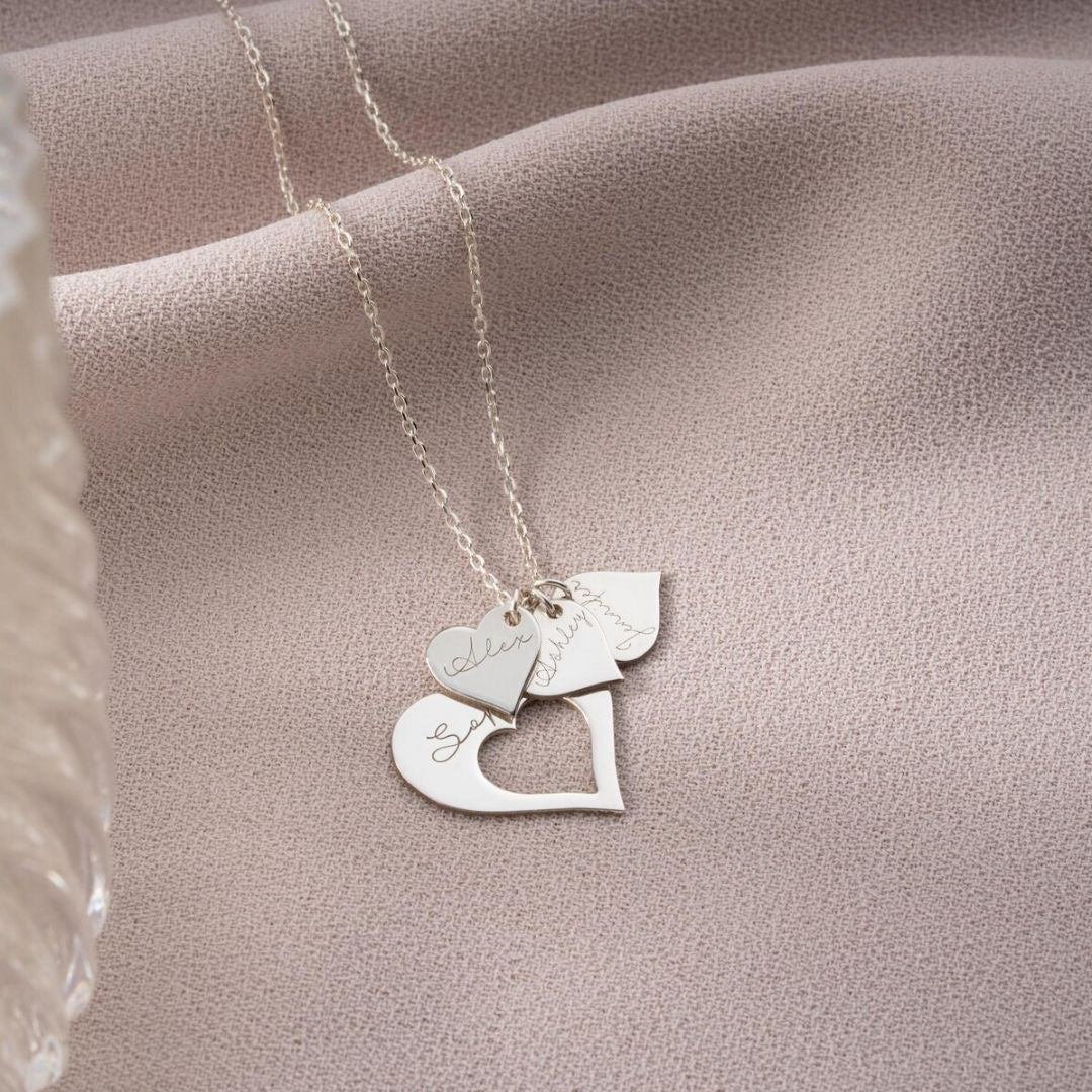 El collar Corazones de familia incluye tres colgantes en forma de corazón con elegantes grabados, expuestos sobre un suave tejido rosa, perfecto como regalo personalizado y lleno de significado.