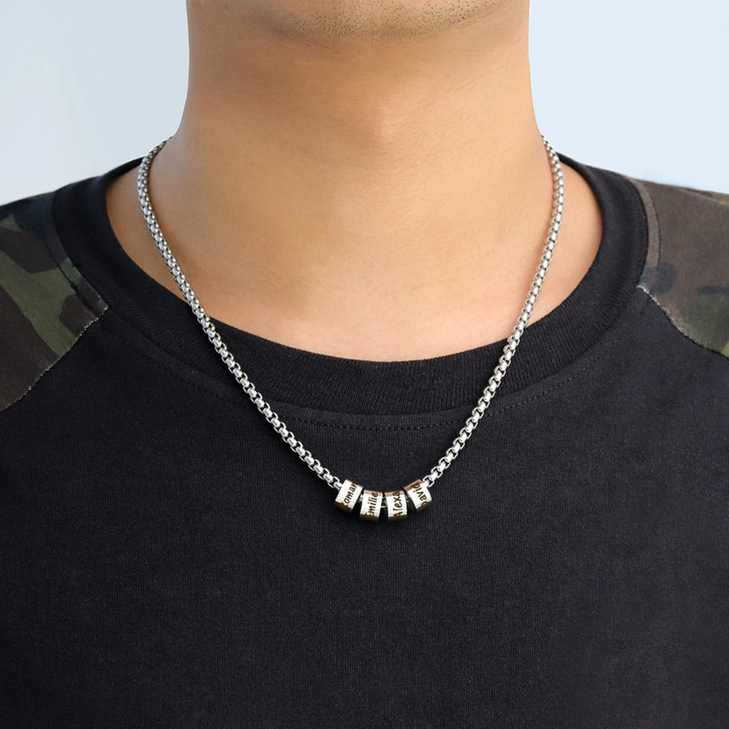 Una persona lleva una camiseta negra con parches de camuflaje en los hombros y un collar de cadena con dijes personalizados con cinco cuentas metálicas con texto grabado.