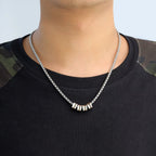 Una persona lleva una camiseta negra con parches de camuflaje en los hombros y un collar de cadena con dijes personalizados con cinco cuentas metálicas con texto grabado.