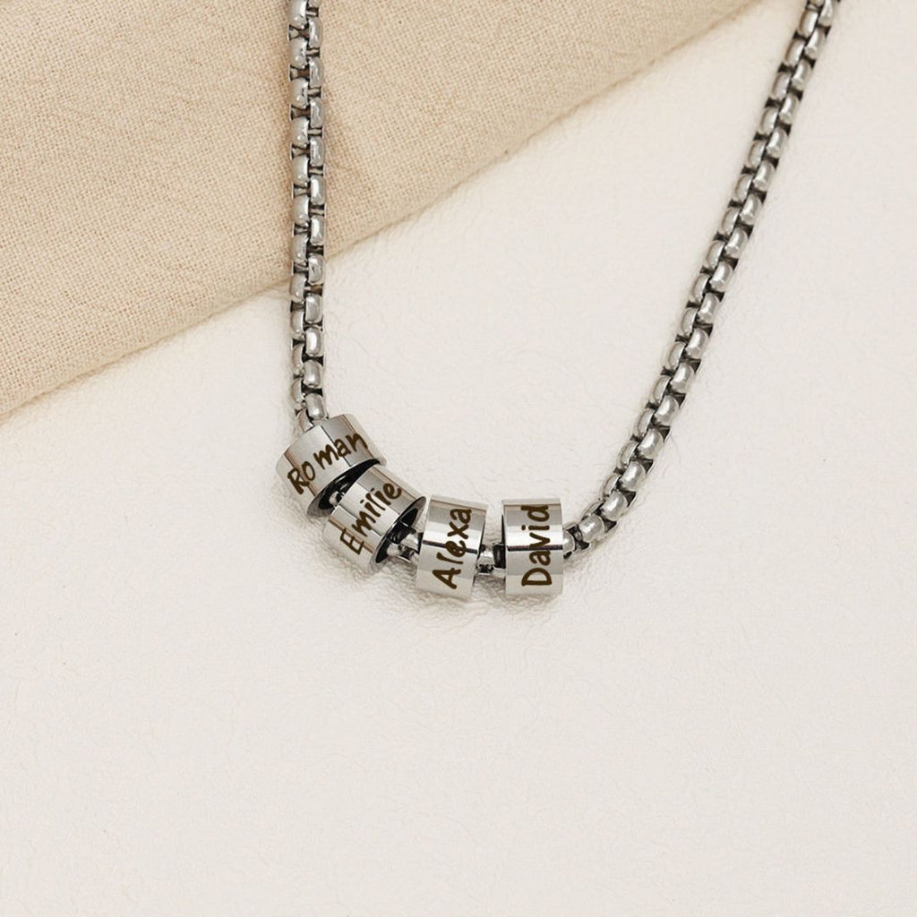 A Collar de cadena con dijes personalizados en plata, con cuatro dijes grabados con los nombres Roman, Emilie, Alexa y David, expuestos sobre una superficie beige clara.