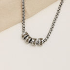A Collar de cadena con dijes personalizados en plata, con cuatro dijes grabados con los nombres Roman, Emilie, Alexa y David, expuestos sobre una superficie beige clara.