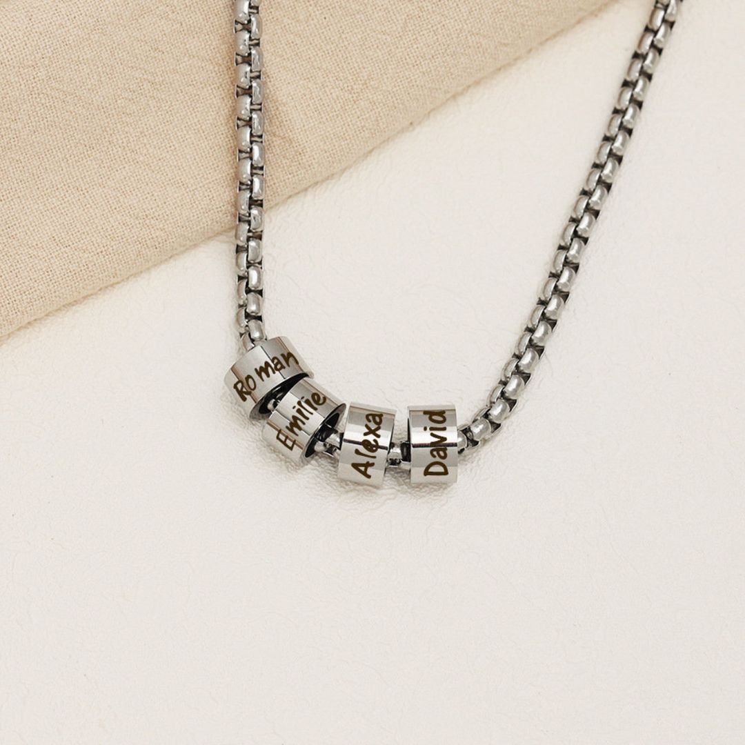A Collar de cadena con dijes personalizados en plata, con cuatro dijes grabados con los nombres Roman, Emilie, Alexa y David, expuestos sobre una superficie beige clara.