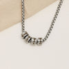 A Collar de cadena con dijes personalizados en plata, con cuatro dijes grabados con los nombres Roman, Emilie, Alexa y David, expuestos sobre una superficie beige clara.