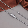 El Collar de cadena con dijes personalizados, con dije cilíndrico segmentado en plata, descansa sobre una superficie gris junto a tela satinada marrón. El broche abierto y los extremos son visibles: un elegante regalo para el Día del Padre.