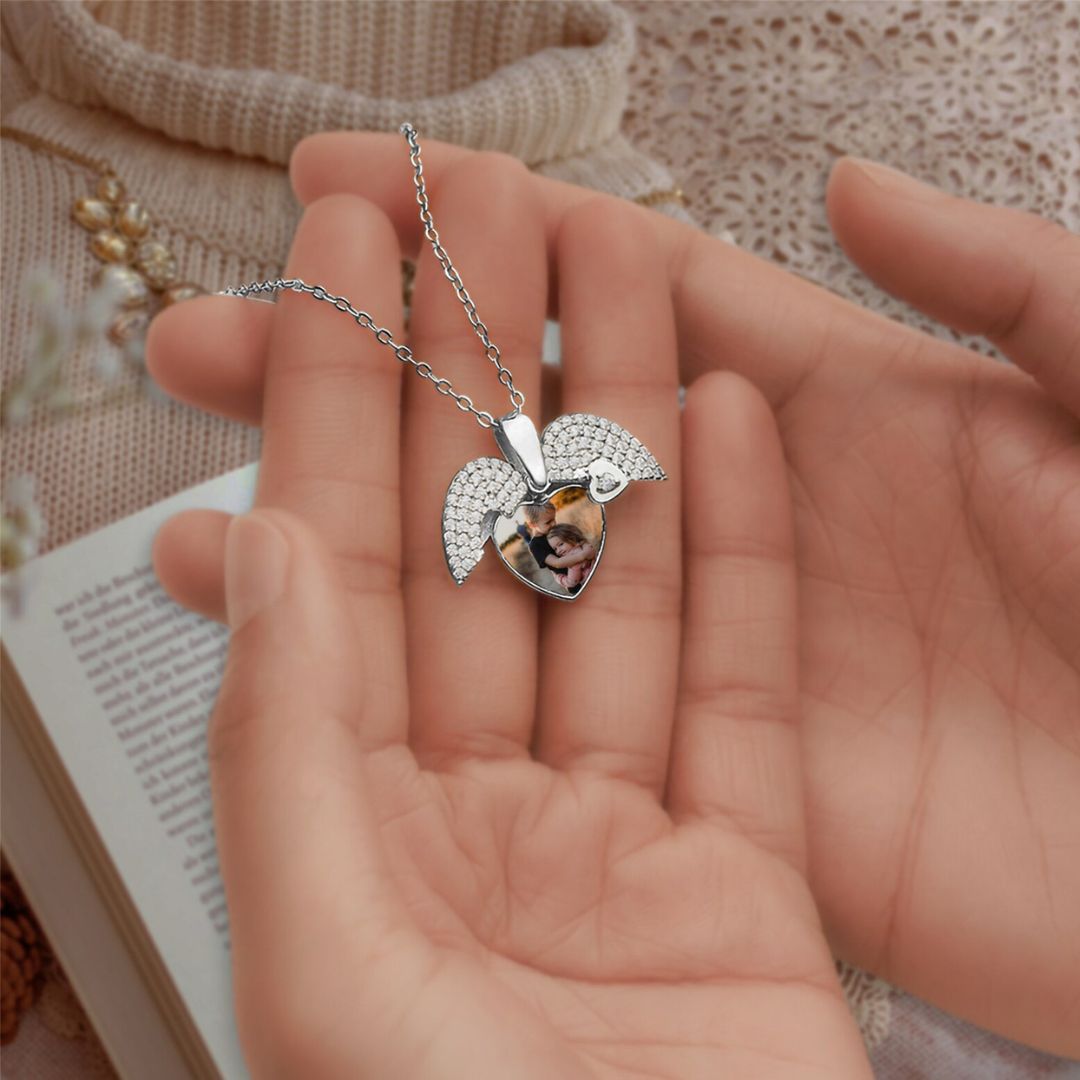 Alguien sostiene el Collar de Corazón Cofre personalizado en color plateado, un medallón en forma de corazón con alas de ángel y una foto personalizable en su interior. Un libro abierto, una tela suave y una luz tenue añaden un toque sentimental a la escena.